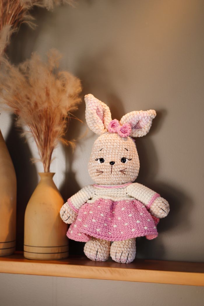 Círculo Amigurumi Pelúcia – Polyester Chenille Yarn (131 m / 85 g) - A crocheted bunny doll wearing a pink dress with white polka dots and a pink bow on its ears, sitting on a wooden shelf next to a vase with dried pampas grass against a neutral gray wall.