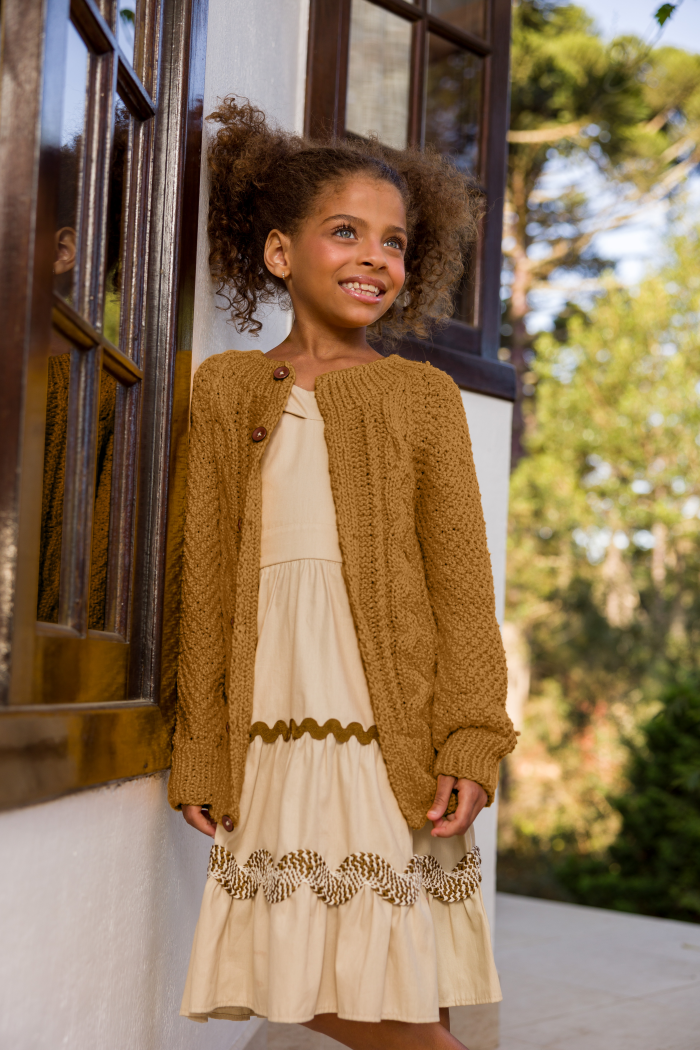Circulo InLove - 100% Brazilian Cotton Yarn (125m/100g) - A young girl with curly hair stands outdoors wearing a cream-colored dress with a textured mustard-brown cardigan.