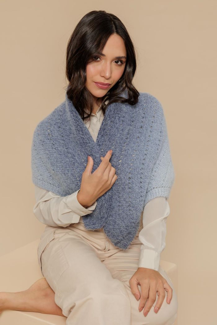 Círculo Marfim – Acrylic Gradient Cake Yarn (400 m / 200 g) - A woman wearing a light blue knit wrap shawl over a white blouse and beige trousers, posed against a solid beige background.