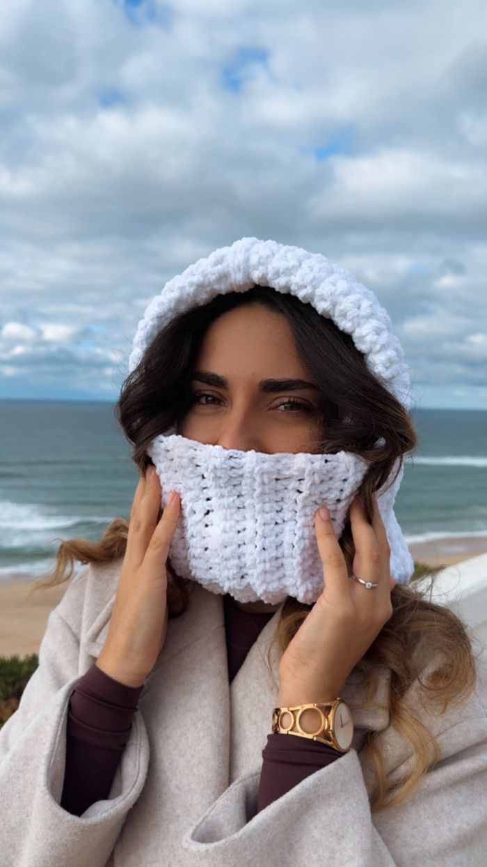 Círculo Velvet – 100% Polyester Chenille Yarn (165 m / 100 g) - A woman wearing a chunky white crocheted beanie and matching face covering stands outdoors by the beach with the ocean and cloudy sky in the background.