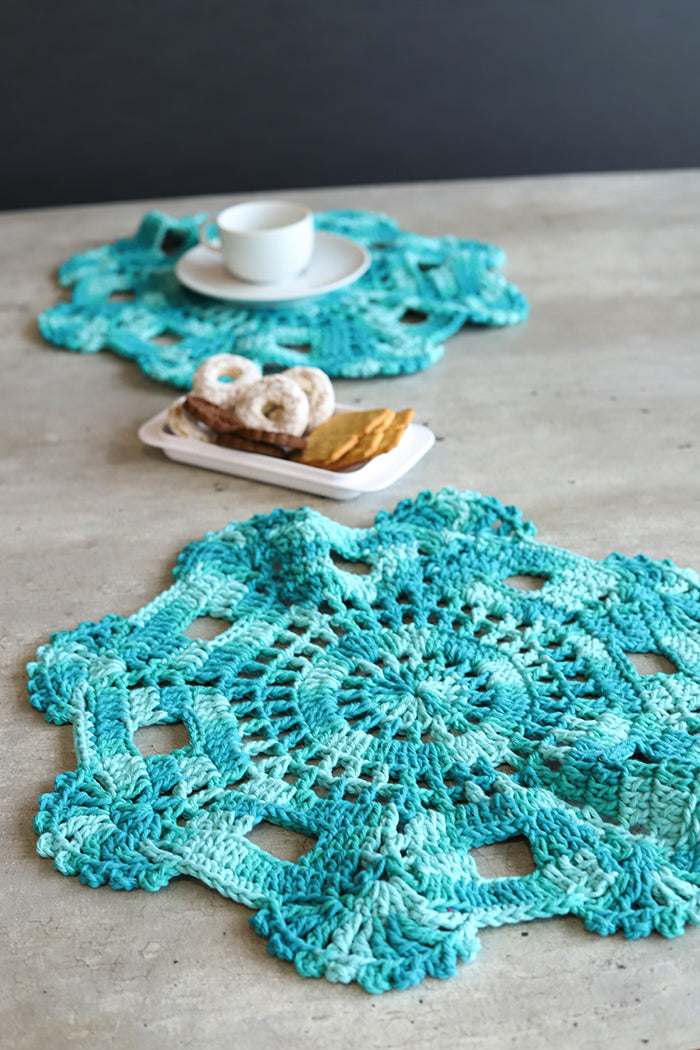 Círculo Natural Cotton Multicolor 4/6 – Cotton Yarn (226 m / 200 g) – Two handmade crocheted doily table mats in a turquoise gradient, styled on a gray concrete tabletop with a white teacup and pastries.