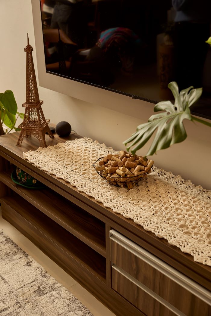 Circulo Barroco Natural Brilho  - Natural Cotton Yarn Gold/Silver, 657m / 433m, 400g - A cream-colored crocheted lace runner tablecloth draped over a dark wood TV stand in a living room setting, styled with a small Eiffel Tower figurine, a bowl of decorative stones, and a potted monstera plant.