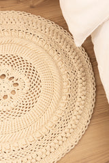 Circulo Barroco Natural Brilho  - Natural Cotton Yarn Gold/Silver, 657m / 433m, 400g - A round, hand-crocheted area rug in a natural beige color, styled on a light wood floor next to a white pillow.