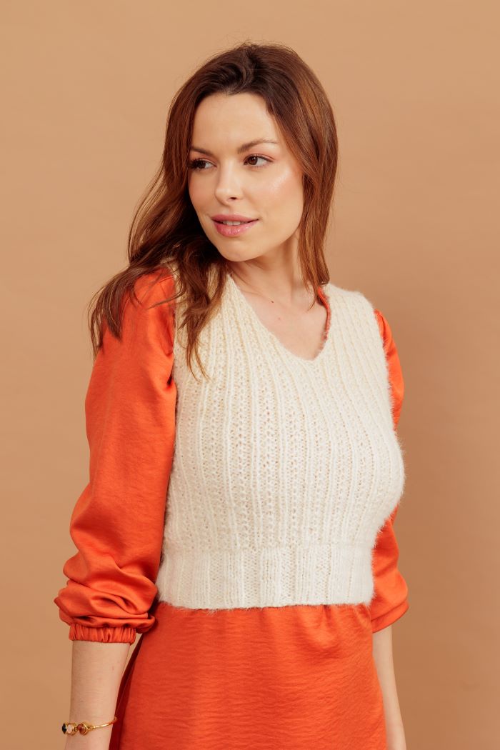 Círculo Marfim – Acrylic Gradient Cake Yarn (400 m / 200 g) - A woman wearing a cream cable-knit V-neck sweater vest over bright orange long-sleeve puff-sleeve top and matching orange skirt, posed against a solid warm tan background.
