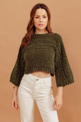 Círculo Velvet – 100% Polyester Chenille Yarn (165 m / 100 g) - A woman wearing a textured olive green cropped knit sweater with three-quarter bell sleeves and white high-waisted pants, standing against a solid warm tan background.