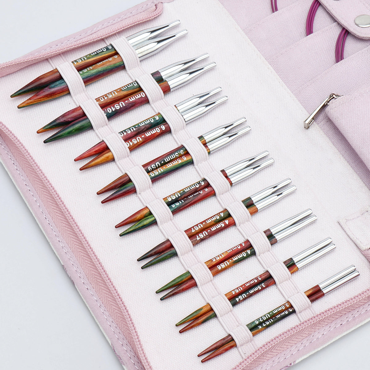 KnitPro Symfonie 2 Special interchangeable needle set, 10 cm, Deluxe - A pink fabric needle case holding multiple knitting needles arranged in elastic loops. The needles have silver metal tips and colored shafts in green, orange, red, and brown tones, with some white text printed on them.